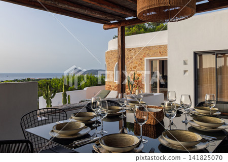 Villa dining terrace on Ibiza, broad table setting beneath a woven lamp and timber roof. Villa dining terrace on Ibiza, broad table setting beneath a woven lamp and timber roof. 141825070