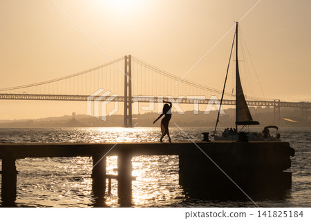 Sunset at the pier with a silhouette of a person walking near the water and a sailboat in the background Sunset at the pier with a silhouette of a person walking near the water and a sailboat in the background 141825184