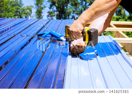 Builder uses power tool to attach metal panels on roof at construction site. Builder uses power tool to attach metal panels on roof at construction site. 141825381