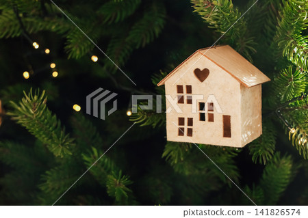 Decorative wooden house ornament hanging on a Christmas tree with warm lights Decorative wooden house ornament hanging on a Christmas tree with warm lights 141826574