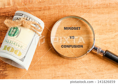 A dollar bill is rolled and tied with a string. A magnifying glass is placed nearby, focusing on the words mindset and confusion written on the wood A dollar bill is rolled and tied with a string. A magnifying glass is placed nearby, focusing on the words mindset and confusion written on the wood 141826859