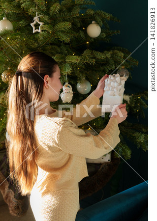 Teen girl holding Christmas gift beside decorated tree at home Teen girl holding Christmas gift beside decorated tree at home 141826913