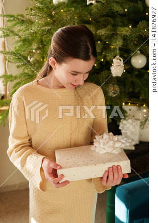 Teen girl looking at Christmas gift with joy beside decorated tree at home Teen girl looking at Christmas gift with joy beside decorated tree at home 141826927