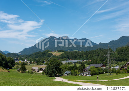 Panoramic view of a green Alpine valley with rural houses, roads, forests and mountain range in the distance. Panoramic view of a green Alpine valley with rural houses, roads, forests and mountain range in the distance. 141827271