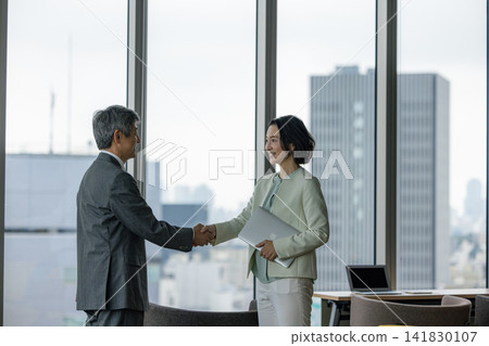 A senior man and a senior woman shake hands in an office by the window. A senior man and a senior woman shake hands in an office by the window. 141830107