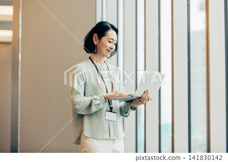 一位五十多歲的女商人站在辦公室窗邊，手裡拿著一台筆記型電腦。 141830142