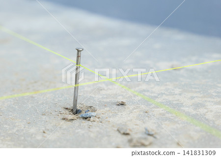 Close-up of a nail hammered into rough concrete with intersecting yellow lines at its base, showcasing precision in construction and alignment for outdoor tiling around the house Close-up of a nail hammered into rough concrete with intersecting yellow lines at its base, showcasing precision in construction and alignment for outdoor tiling around the house 141831309