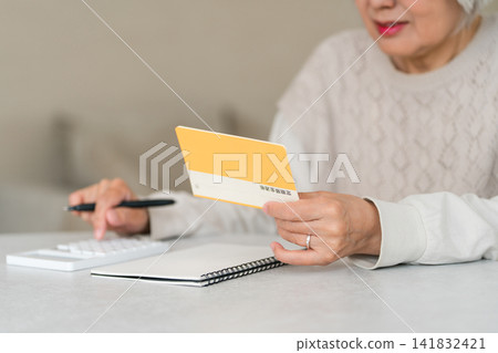 Senior woman calculating using a calculator while looking at her savings passbook (retirement funds, living expenses, pension) 141832421