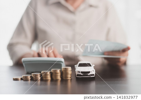 Person reviewing financial papers with a calculator beside a white model car and coin stacks, representing auto loan comparison, vehicle budgeting, purchase planning, and ownership costs. Person reviewing financial papers with a calculator beside a white model car and coin stacks, representing auto loan comparison, vehicle budgeting, purchase planning, and ownership costs. 141832977