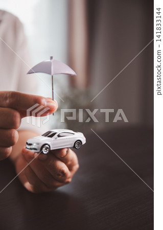 Hands hold a white car model beneath a small umbrella, symbolizing vehicle insurance, accident protection, risk coverage, financial security, responsible ownership, and safe mobility planning. 141833144