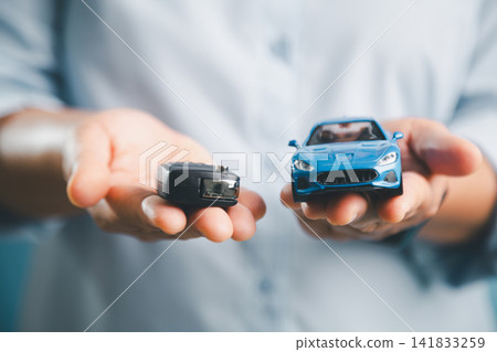 Person holding a car key and blue toy car, representing vehicle ownership, car insurance, auto loan approval, rental service, leasing, and automobile handover concept. Person holding a car key and blue toy car, representing vehicle ownership, car insurance, auto loan approval, rental service, leasing, and automobile handover concept. 141833259