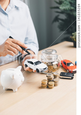 Consultant holding white toy car with piggy bank, coin jar, toy cars, coins, and car key, representing car finance consultation, savings, and purchase planning. 141833284