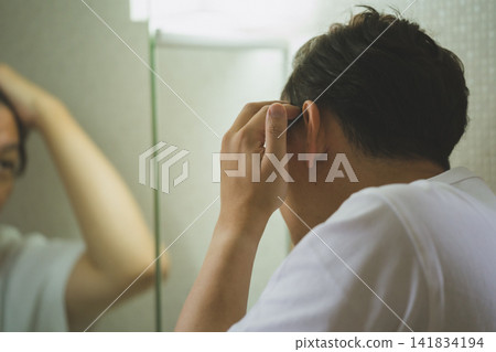 A man in his 40s checks his thinning hair by touching the side of his hair at the sink, viewed from behind. A man in his 40s checks his thinning hair by touching the side of his hair at the sink, viewed from behind. 141834194