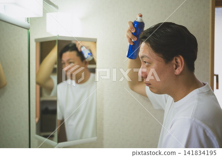 A 40-year-old man performs scalp care by spraying hair growth treatment on his head in front of the bathroom mirror. A 40-year-old man performs scalp care by spraying hair growth treatment on his head in front of the bathroom mirror. 141834195