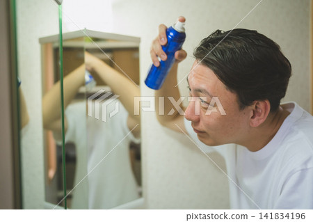 一位40多歲的男士在浴室鏡子前使用噴霧進行頭髮護理。 141834196