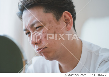 一位 40 歲的男子皺著眉頭對著桌面上的鏡子,為臉頰上的瑕疵而煩惱,並討論著護膚。 一位 40 歲的男子皺著眉頭對著桌面上的鏡子,為臉頰上的瑕疵而煩惱,並討論著護膚。 141836677