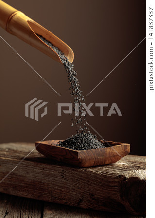 Grains of black sesame are poured with a wooden spoon in the wooden dish. Grains of black sesame are poured with a wooden spoon in the wooden dish. 141837337