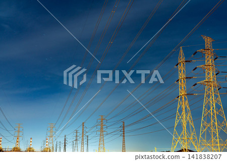 High voltage electricity transmission towers and power lines under a blue night sky with stars near an electrical substation. Modern energy infrastructure, utility networks and power distribution. High voltage electricity transmission towers and power lines under a blue night sky with stars near an electrical substation. Modern energy infrastructure, utility networks and power distribution. 141838207