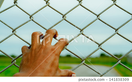 Man hand holding metal chain link fence. Refugee and immigrant concept. Life and freedom. Anguish, gloom, and persecution feeling. Man hand grabbing cage or camp fence. Prison fence. Barrier border. Man hand holding metal chain link fence. Refugee and immigrant concept. Life and freedom. Anguish, gloom, and persecution feeling. Man hand grabbing cage or camp fence. Prison fence. Barrier border. 141838309