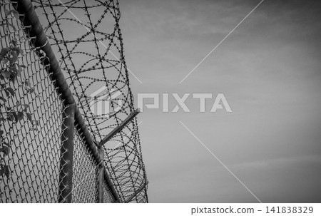Black and white razor wire security fence surrounding a restricted area with dramatic copy space, representing prison security, border protection, restricted access, surveillance and freedom concepts. 141838329