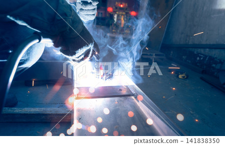 Welder welding metal with argon arc welding machine and has welding sparks and smoke. Man wear protective gloves. Safety in industrial workplace. Welder working with safety. Steel industry technology. Welder welding metal with argon arc welding machine and has welding sparks and smoke. Man wear protective gloves. Safety in industrial workplace. Welder working with safety. Steel industry technology. 141838350
