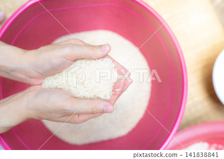 Top view woman hand holding rice above red plastic bowl. Global food crisis. Raw dry rice. Uncooked milled white rice. Zakat and charity concept. Organic cereal grain. Rice price in world market. Top view woman hand holding rice above red plastic bowl. Global food crisis. Raw dry rice. Uncooked milled white rice. Zakat and charity concept. Organic cereal grain. Rice price in world market. 141838841