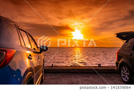 Two modern cars parked beside the sea during a vibrant golden sunset with dramatic clouds and glowing reflections. Coastal road trip adventure, travel freedom, vacation lifestyle and transportation. Two modern cars parked beside the sea during a vibrant golden sunset with dramatic clouds and glowing reflections. Coastal road trip adventure, travel freedom, vacation lifestyle and transportation. 141839064