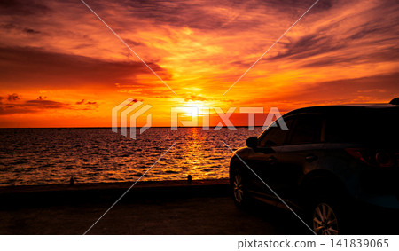 Modern sport utility vehicle parked beside the ocean during a dramatic orange sunset with colorful clouds and glowing reflections. Coastal road trip adventure, travel freedom and transportation. Modern sport utility vehicle parked beside the ocean during a dramatic orange sunset with colorful clouds and glowing reflections. Coastal road trip adventure, travel freedom and transportation. 141839065