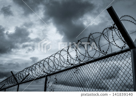 Razor wire security fence beneath dramatic storm clouds, concept of border security, military defense, prison protection, restricted access, surveillance systems and critical infrastructure safety. Razor wire security fence beneath dramatic storm clouds, concept of border security, military defense, prison protection, restricted access, surveillance systems and critical infrastructure safety. 141839140