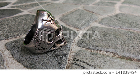 Side view of a silver Gothic-style skull ring against a stone surface background. Side view of a silver Gothic-style skull ring against a stone surface background. 141839454