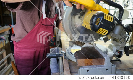 A man in maroon overalls works at a workbench. He lowers a DeWalt circular miter saw onto a block of wood. The yellow and black tool is clamped to the stand. The craftsman carefully cuts the workpiece 141840198