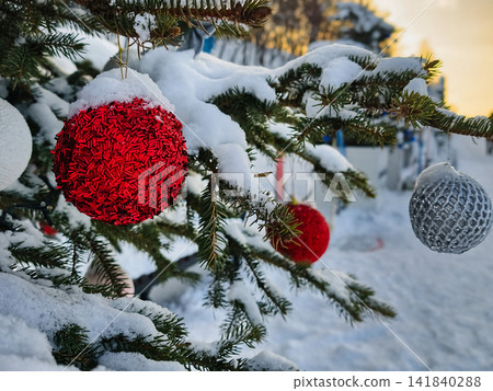 A Christmas tree stands decorated with red and silver ornaments. Snow covers the branches as people celebrate New Year outdoors in a winter scene A Christmas tree stands decorated with red and silver ornaments. Snow covers the branches as people celebrate New Year outdoors in a winter scene 141840288