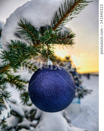 The Christmas tree is covered in snow and has a shiny ornament hanging from a branch. The sun sets in the background, marking the holiday season The Christmas tree is covered in snow and has a shiny ornament hanging from a branch. The sun sets in the background, marking the holiday season 141840323