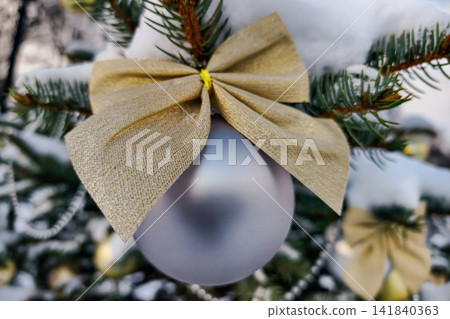 A natural Christmas tree stands outside in winter with decorations. A silver ornament hangs from a branch secured by a golden bow A natural Christmas tree stands outside in winter with decorations. A silver ornament hangs from a branch secured by a golden bow 141840363