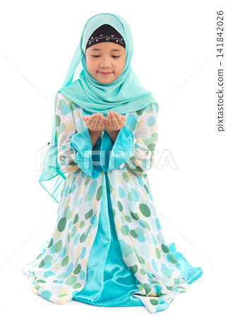 Charming young Asian Muslim child in traditional headscarf kneeling politely holding hands up in prayer looking down. Charming young Asian Muslim child in traditional headscarf kneeling politely holding hands up in prayer looking down. 141842026