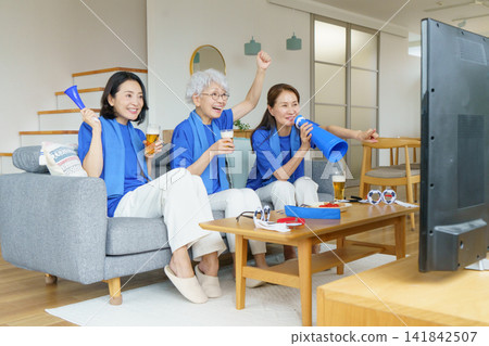 Three middle-aged Japanese women are sitting side-by-side on a living room sofa, watching the Japanese national soccer team on TV. Three middle-aged Japanese women are sitting side-by-side on a living room sofa, watching the Japanese national soccer team on TV. 141842507