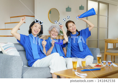 Three middle-aged Japanese women are sitting side-by-side on a living room sofa, watching the Japanese national soccer team on TV. Three middle-aged Japanese women are sitting side-by-side on a living room sofa, watching the Japanese national soccer team on TV. 141842538