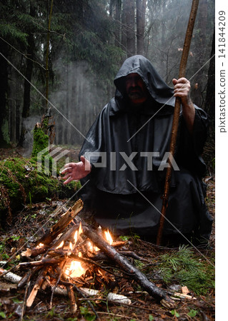 A monk in a black robe sits in the forest by the fire and warms his hands. Mysticism and magic, sorcerers and witches. A monk in a black robe sits in the forest by the fire and warms his hands. Mysticism and magic, sorcerers and witches. 141844209