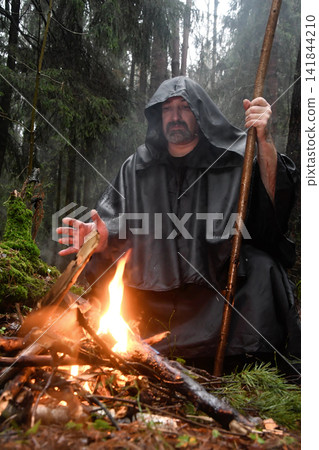A monk in a black robe sits in the forest by the fire and warms his hands. Mysticism and magic, sorcerers and witches. A monk in a black robe sits in the forest by the fire and warms his hands. Mysticism and magic, sorcerers and witches. 141844210