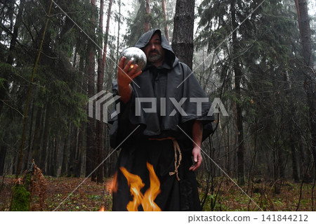 A black-robed monk sits by a fire in the forest and looks into a divination ball. Mysticism and magic, sorcerers and witches A black-robed monk sits by a fire in the forest and looks into a divination ball. Mysticism and magic, sorcerers and witches 141844212