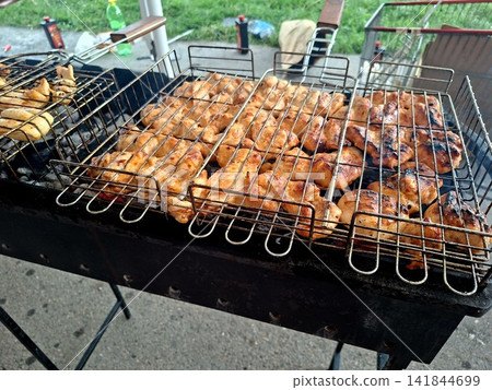 Delicious chicken meat cooking in wire grate mesh over hot barbecue grill charcoal embers for summer picnic outdoor dinner party food preparation Delicious chicken meat cooking in wire grate mesh over hot barbecue grill charcoal embers for summer picnic outdoor dinner party food preparation 141844699