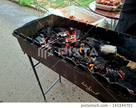 Side angle view of outdoor metal barbecue mangal filled with glowing charcoal embers ready for grilling meat during summer picnic family gathering Side angle view of outdoor metal barbecue mangal filled with glowing charcoal embers ready for grilling meat during summer picnic family gathering 141844701