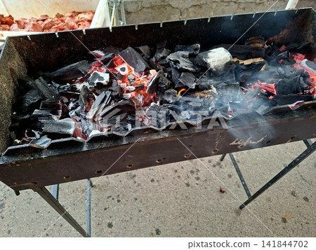 Close up of burning charcoal embers with smoke rising from metal barbecue grill prepared for outdoor picnic cooking and summer food grilling party Close up of burning charcoal embers with smoke rising from metal barbecue grill prepared for outdoor picnic cooking and summer food grilling party 141844702