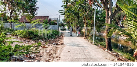Riverside village path with pedestrians and shady trees Riverside village path with pedestrians and shady trees 141844836