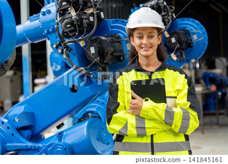 Young female engineer in high-visibility gear smiles confidently beside large blue robotic arm in industrial setting Young female engineer in high-visibility gear smiles confidently beside large blue robotic arm in industrial setting 141845161