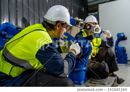 Two technicians in safety gear collaborate on blue robotic arm with focused expressions in industrial workshop Two technicians in safety gear collaborate on blue robotic arm with focused expressions in industrial workshop 141845164