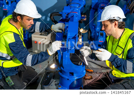 Two technicians in high-visibility vests meticulously repair blue robotic arm in industrial workshop with focused expressions Two technicians in high-visibility vests meticulously repair blue robotic arm in industrial workshop with focused expressions 141845167
