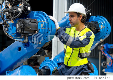 Male technician in safety gear inspects blue robotic arm with focused expression in industrial workshop Male technician in safety gear inspects blue robotic arm with focused expression in industrial workshop 141845168
