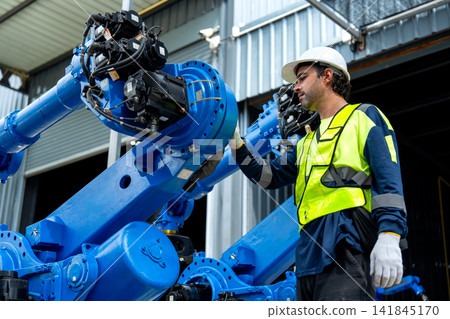 Male technician in safety gear inspects blue robotic arm with focused expression in industrial workshop 141845170