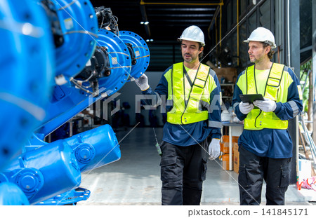 Two technicians in high-visibility vests inspect blue robotic arm in industrial workshop with focused expressions Two technicians in high-visibility vests inspect blue robotic arm in industrial workshop with focused expressions 141845171